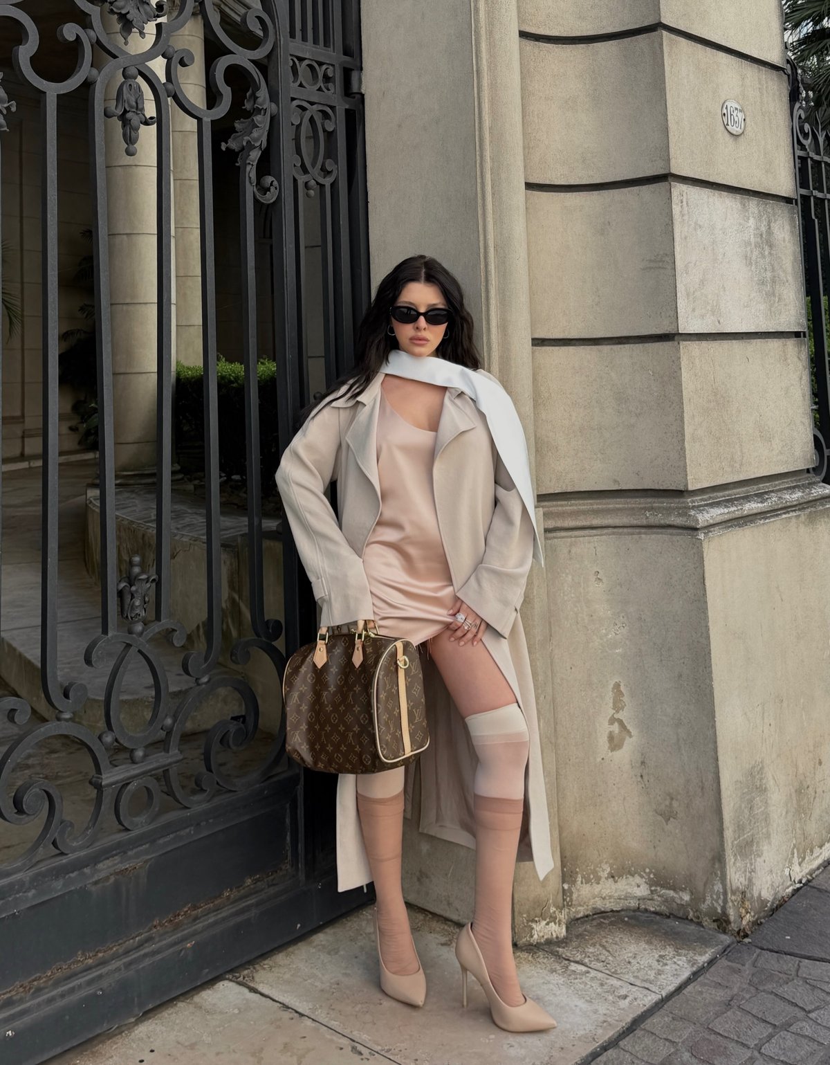Model in a blush slip dress, beige trench and white scarf with a Louis Vuitton bag by a wrought-iron gate
