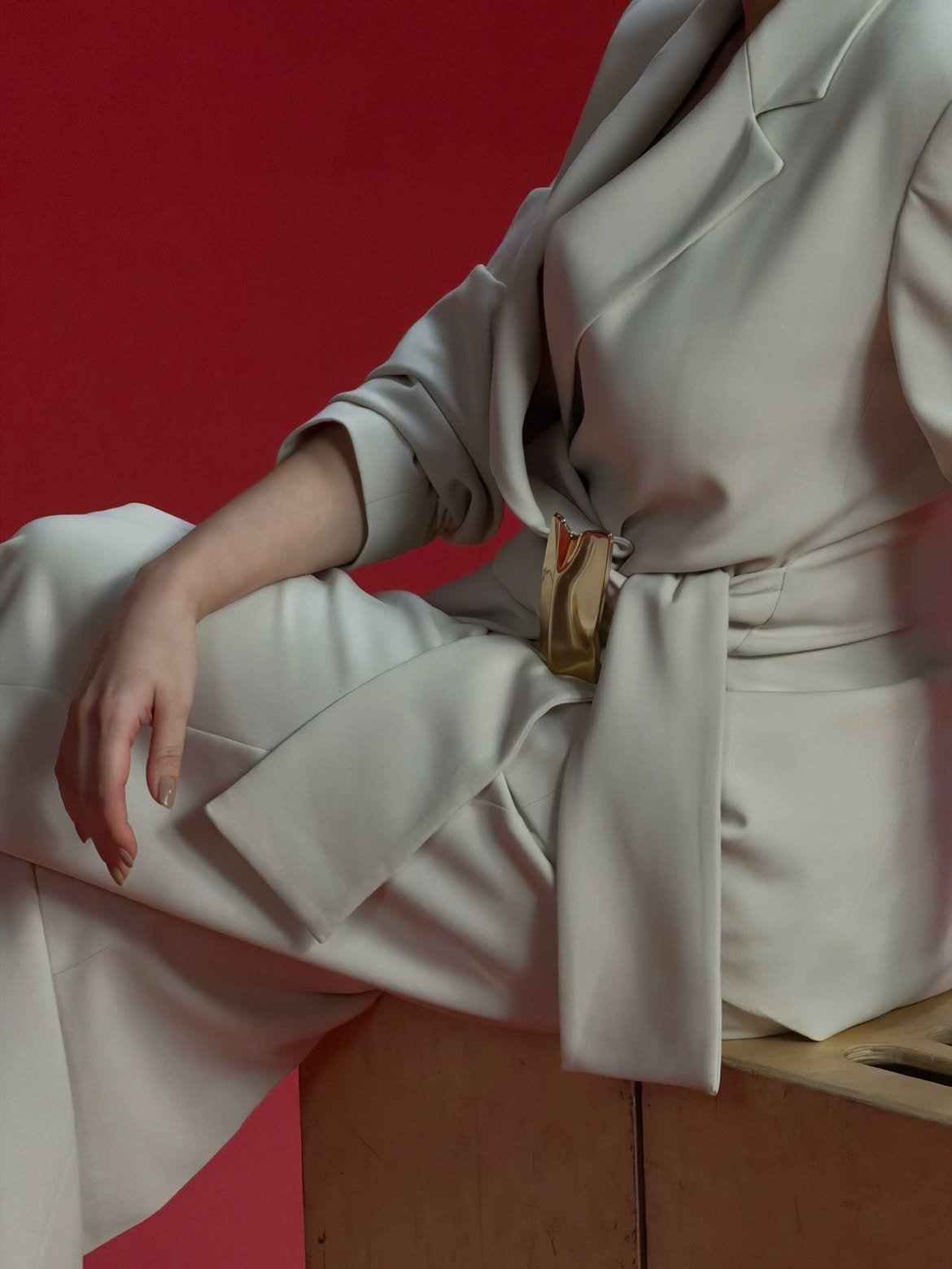 Cream belted suit with a sculptural gold buckle against a red wall