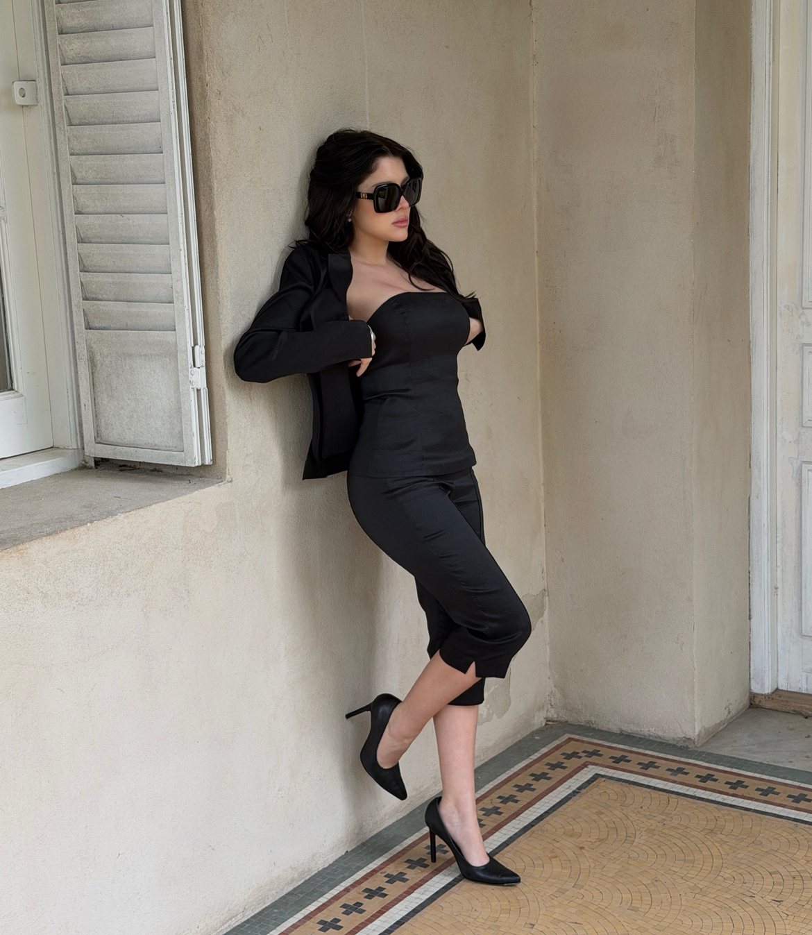 Model in a black strapless top, capri trousers and sunglasses leaning by a shuttered window