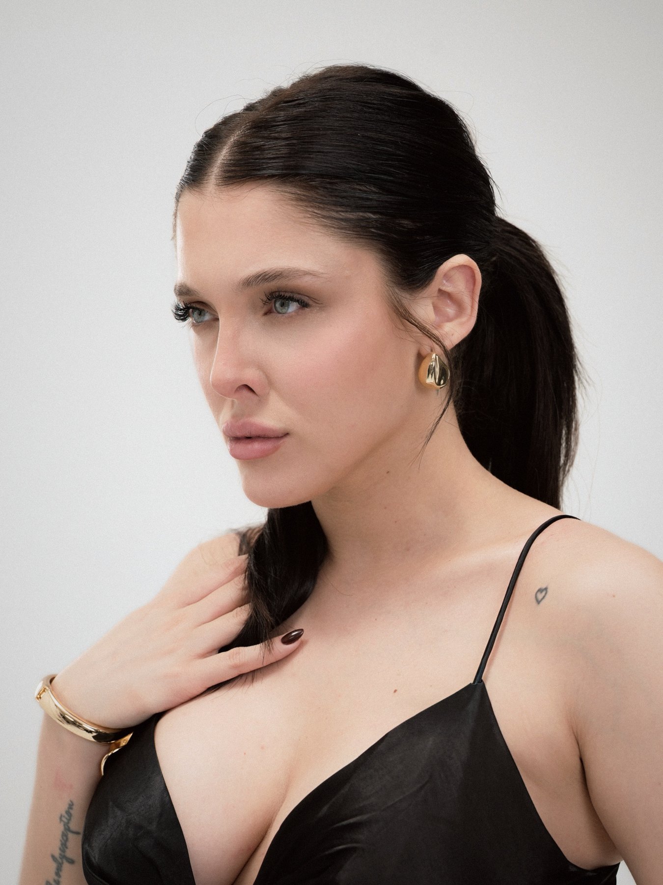Close portrait of Sofía Gonet in black satin, gold earring and cuff, hand at her collarbone